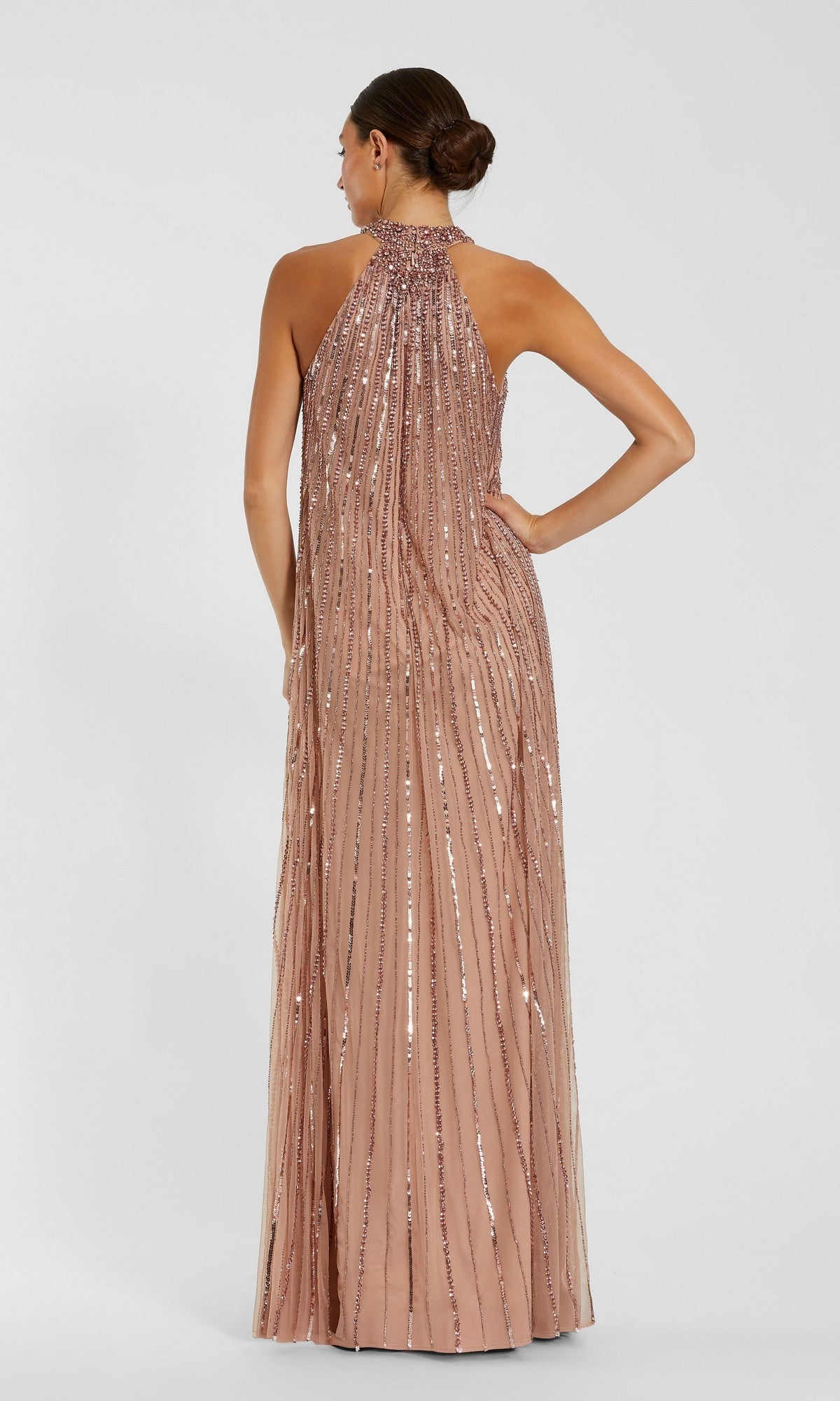 A woman stands with her back to the camera wearing the Mac Duggal 23043 long formal dress in blush, featuring vertical sequin embellishments, a high neckline, and floor-length embellished mesh.
